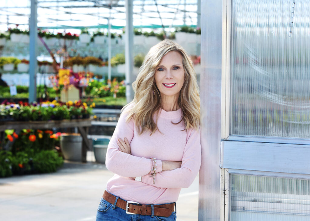 Woman in a pink sweater leaning on a greenhouse door with the green house in the background