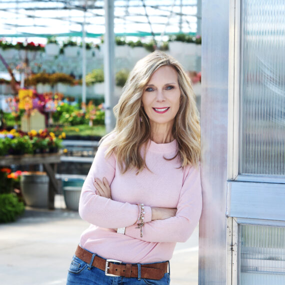 Woman in a pink sweater leaning on a greenhouse door with the green house in the background