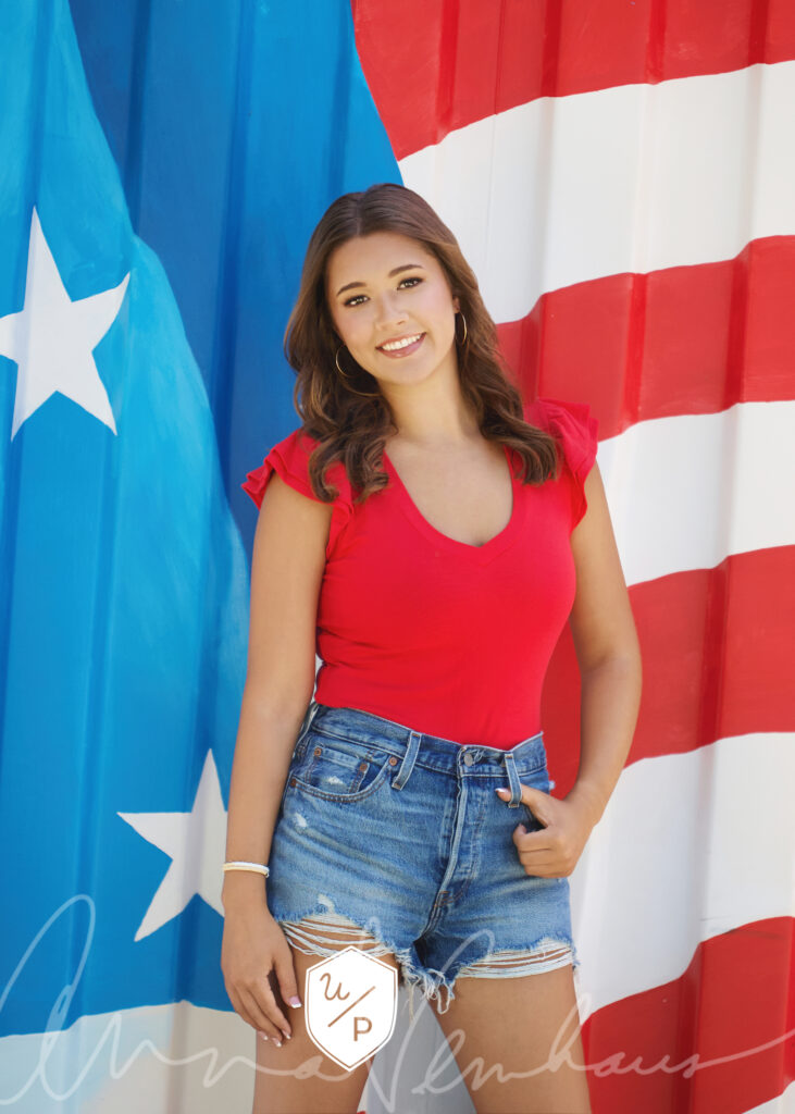 brunette senior girl in red top and denim shorts in front of American flag mural