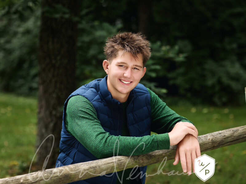 senior boy in forest green shirt and navy vest posed leaning on fence post